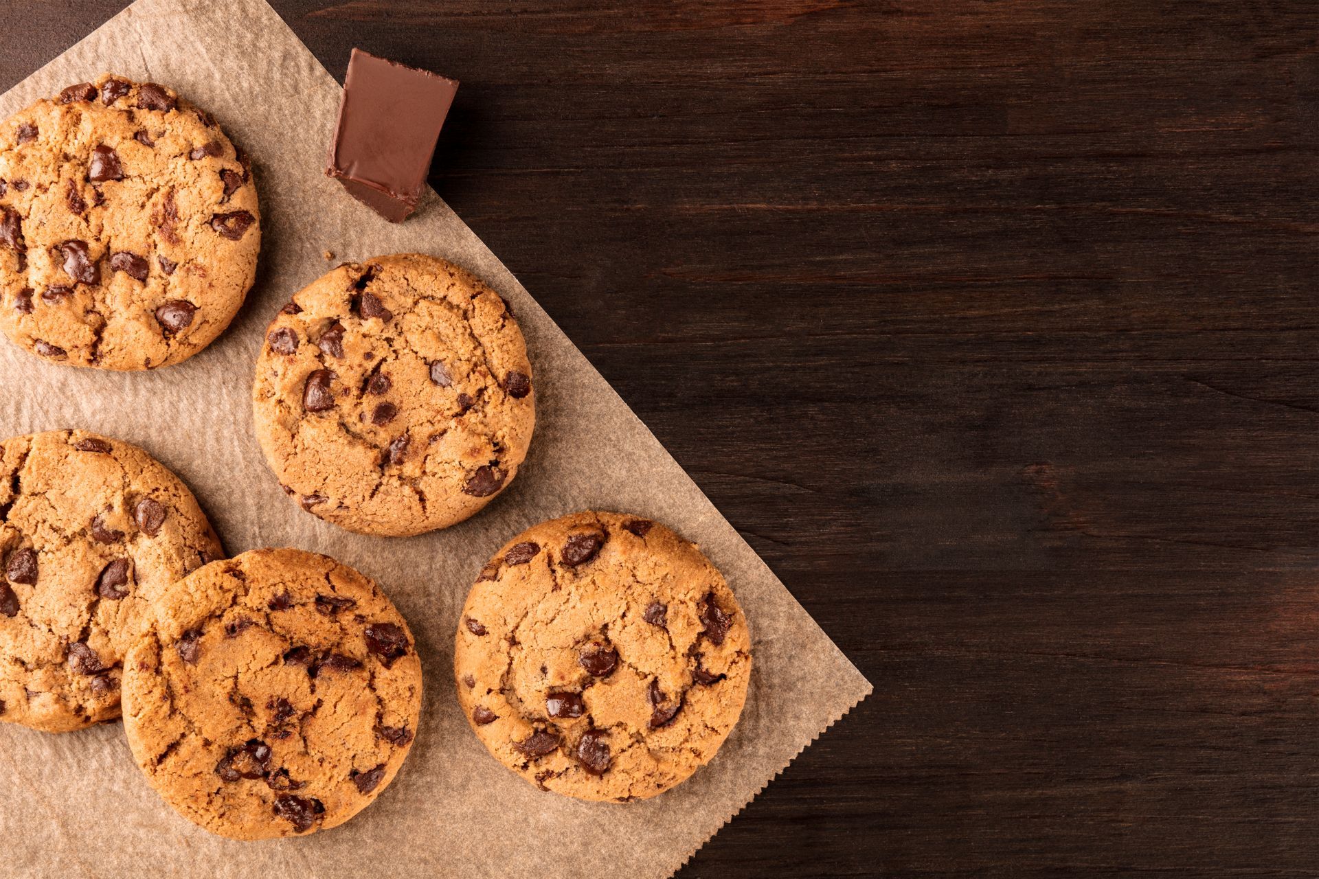Des biscuits aux pépites de chocolat sur une serviette brune, avec un petit morceau de chocolat sur une surface en bois foncé.