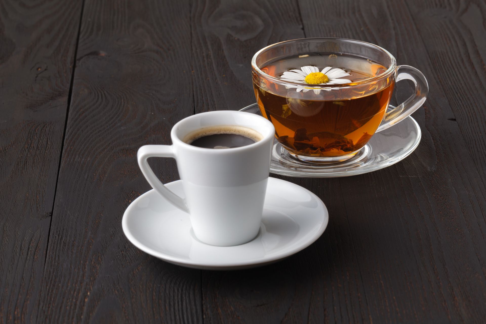 Tasse à café blanche sur sa soucoupe, à côté d'une tasse à thé en verre avec un citron, sur une table en bois foncé.