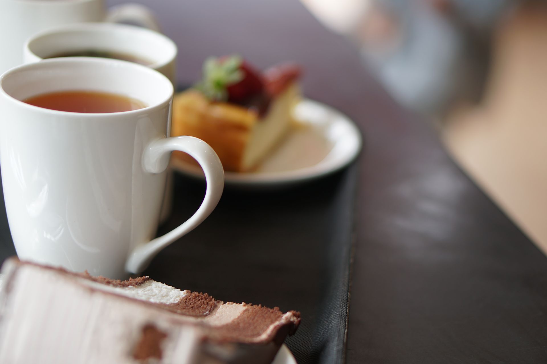 Une tasse de thé blanche sur une table, avec une assiette à dessert floue en arrière-plan.