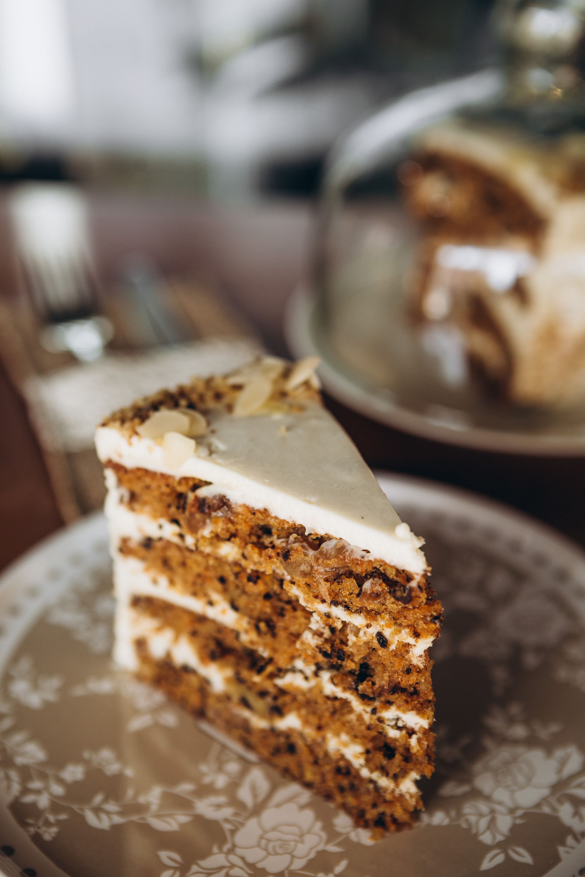 Une part de gâteau aux carottes à étages, nappée de glaçage blanc, présentée sur une assiette à motifs, avec un gâteau flou en arrière-plan.