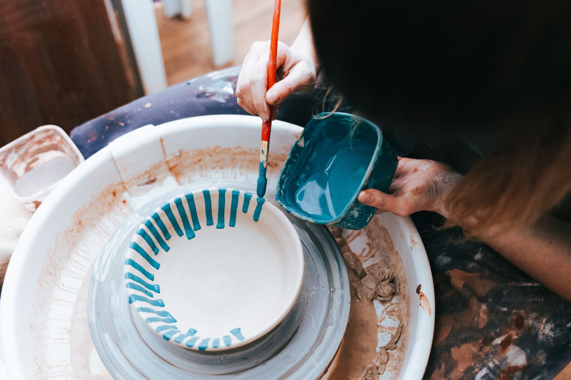 Des mains peignent des rayures turquoise sur une assiette blanche à côté d'une tasse bleue sur une table de travail en désordre