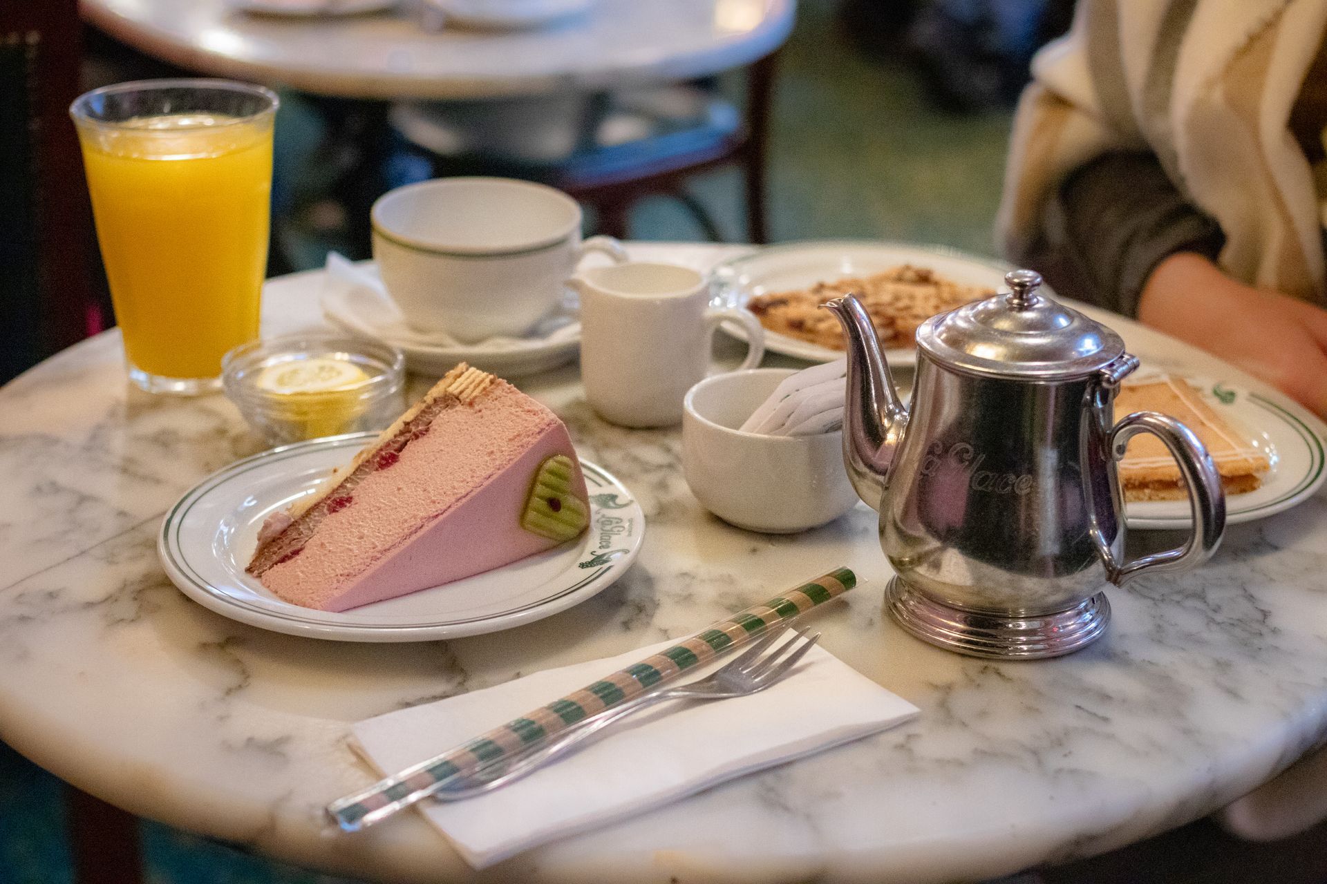 Service à thé avec part de gâteau, jus d'orange et théière en argent sur une table de café en marbre.