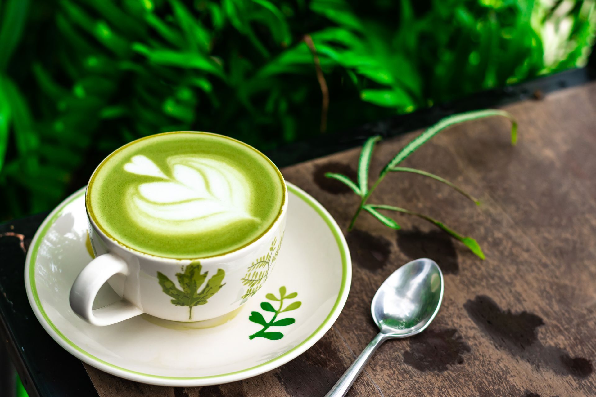 Un matcha latte vert servi dans une tasse et une soucoupe blanches, à côté d'une cuillère, sur une table rustique, avec des feuilles vertes à proximité.