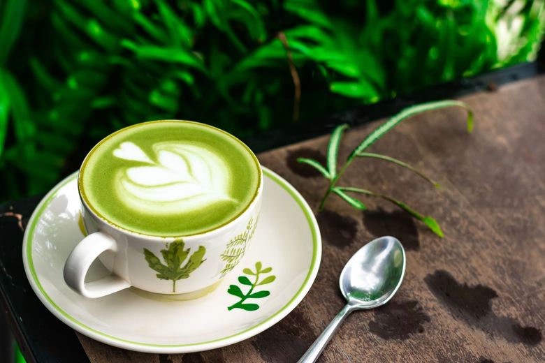 Un matcha latte vert servi dans une tasse et une soucoupe blanches, à côté d'une cuillère, sur une table rustique, avec des feuilles vertes à proximité.