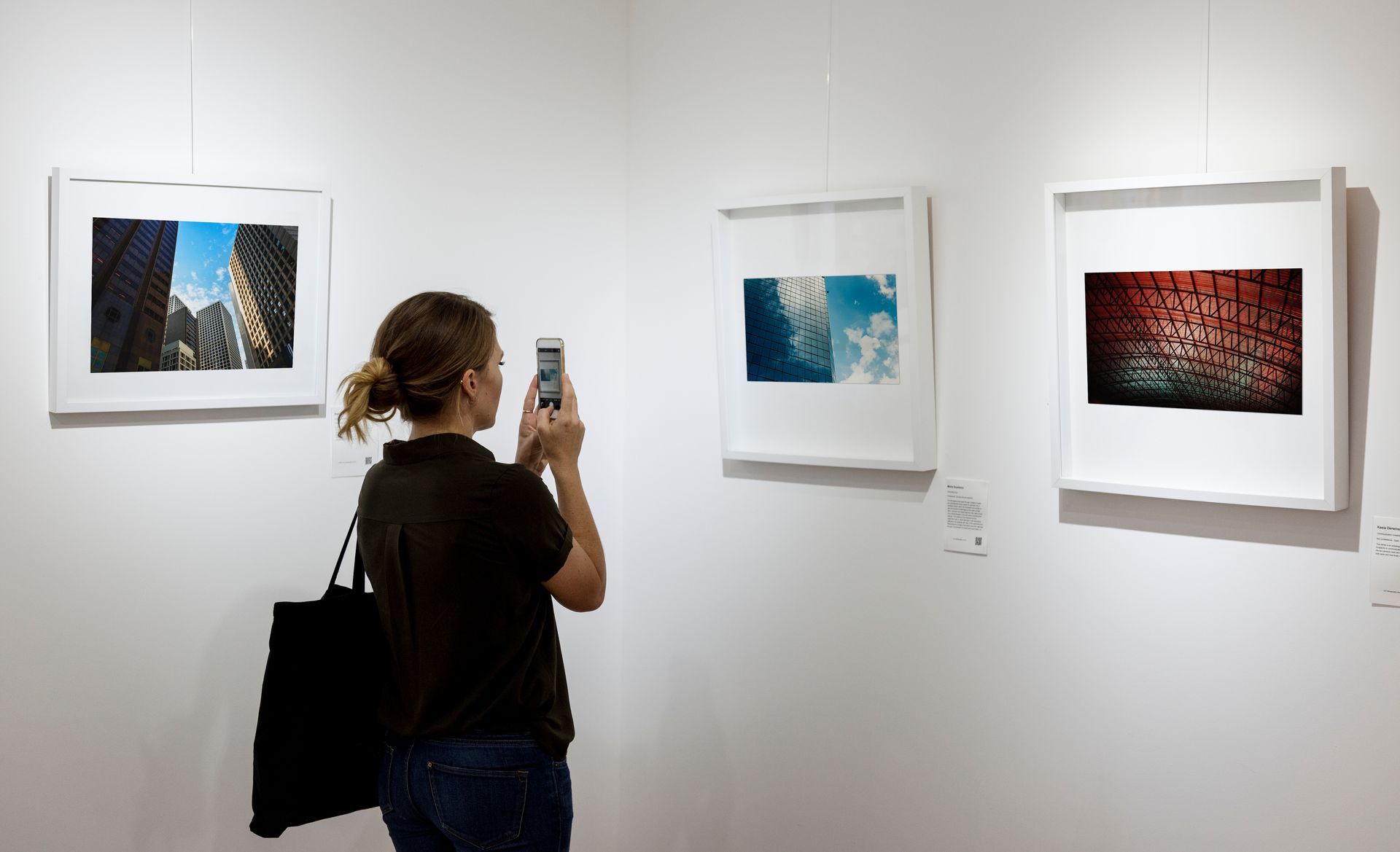 Une personne photographie trois œuvres d'art encadrées accrochées sur un mur blanc de galerie.