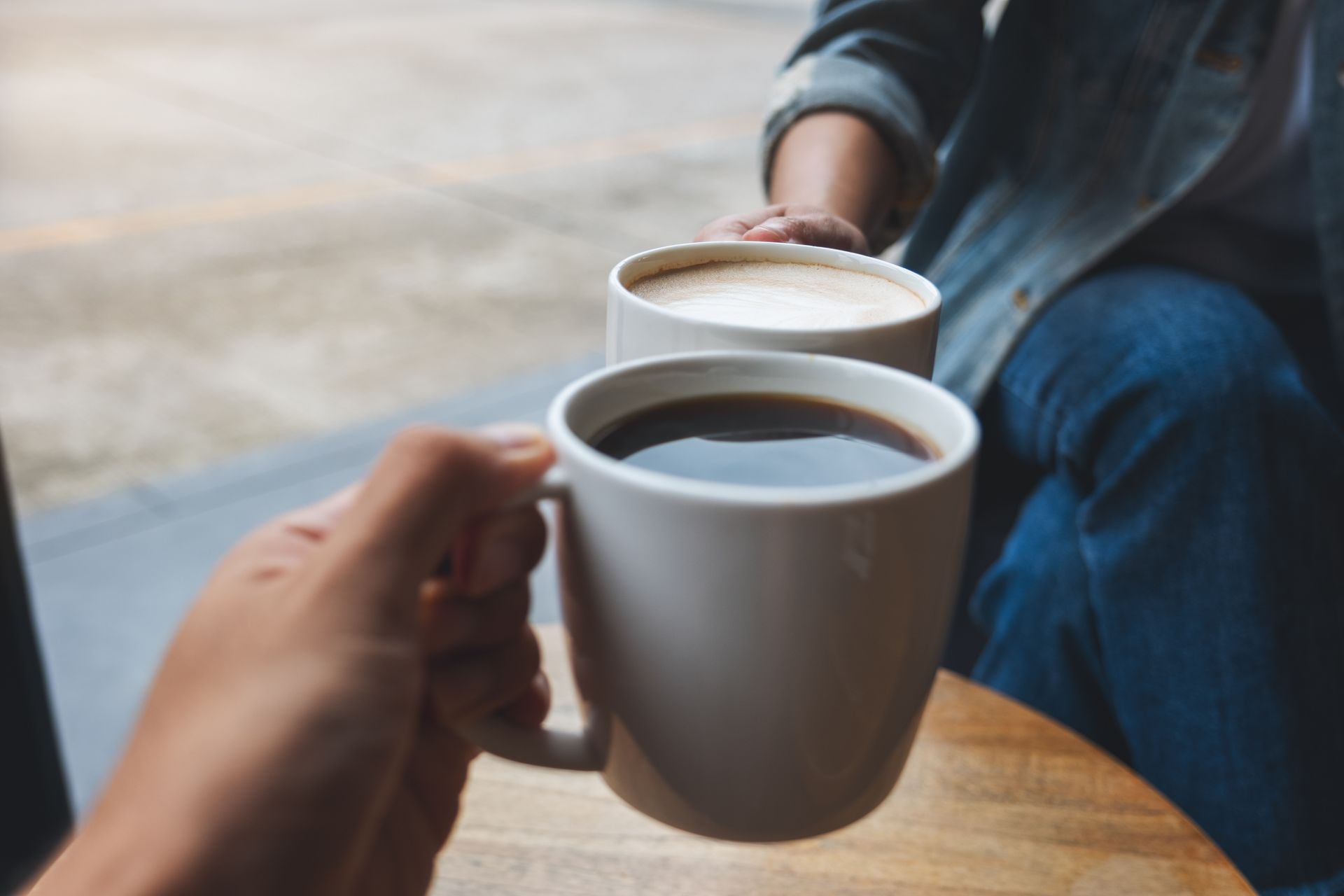 Deux personnes trinquent avec des tasses à café blanches à une table, avec un bras vêtu de denim en arrière-plan.