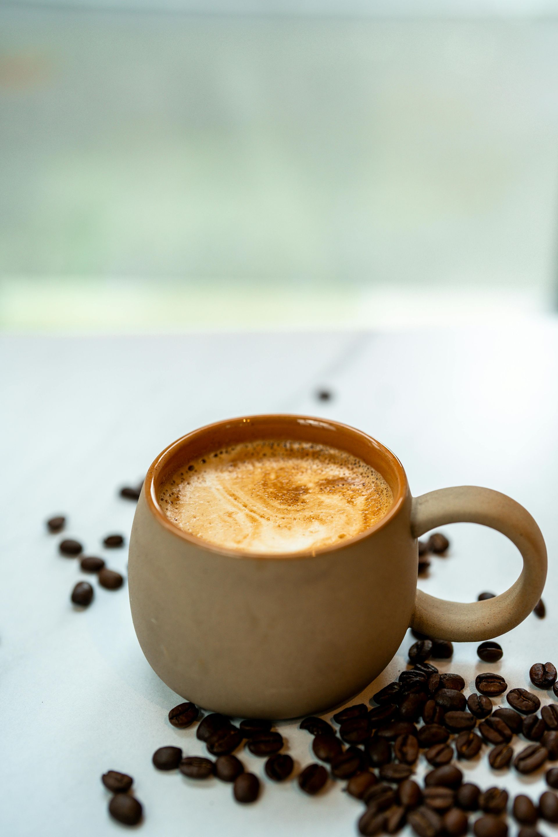 Mug de café brun avec de la mousse, entouré de grains de café éparpillés sur une surface blanche.