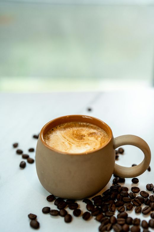 Mug de café brun avec de la mousse, entouré de grains de café éparpillés sur une surface blanche.