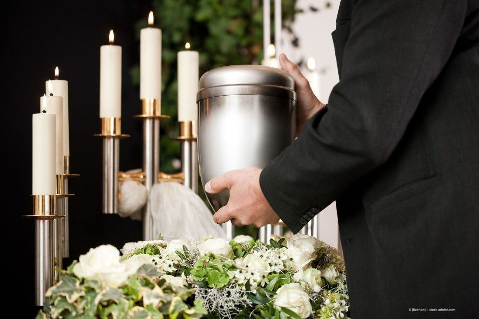 Person in schwarzem Anzug hält eine silberfarbene Urne vor brennenden Kerzen und Blumenschmuck