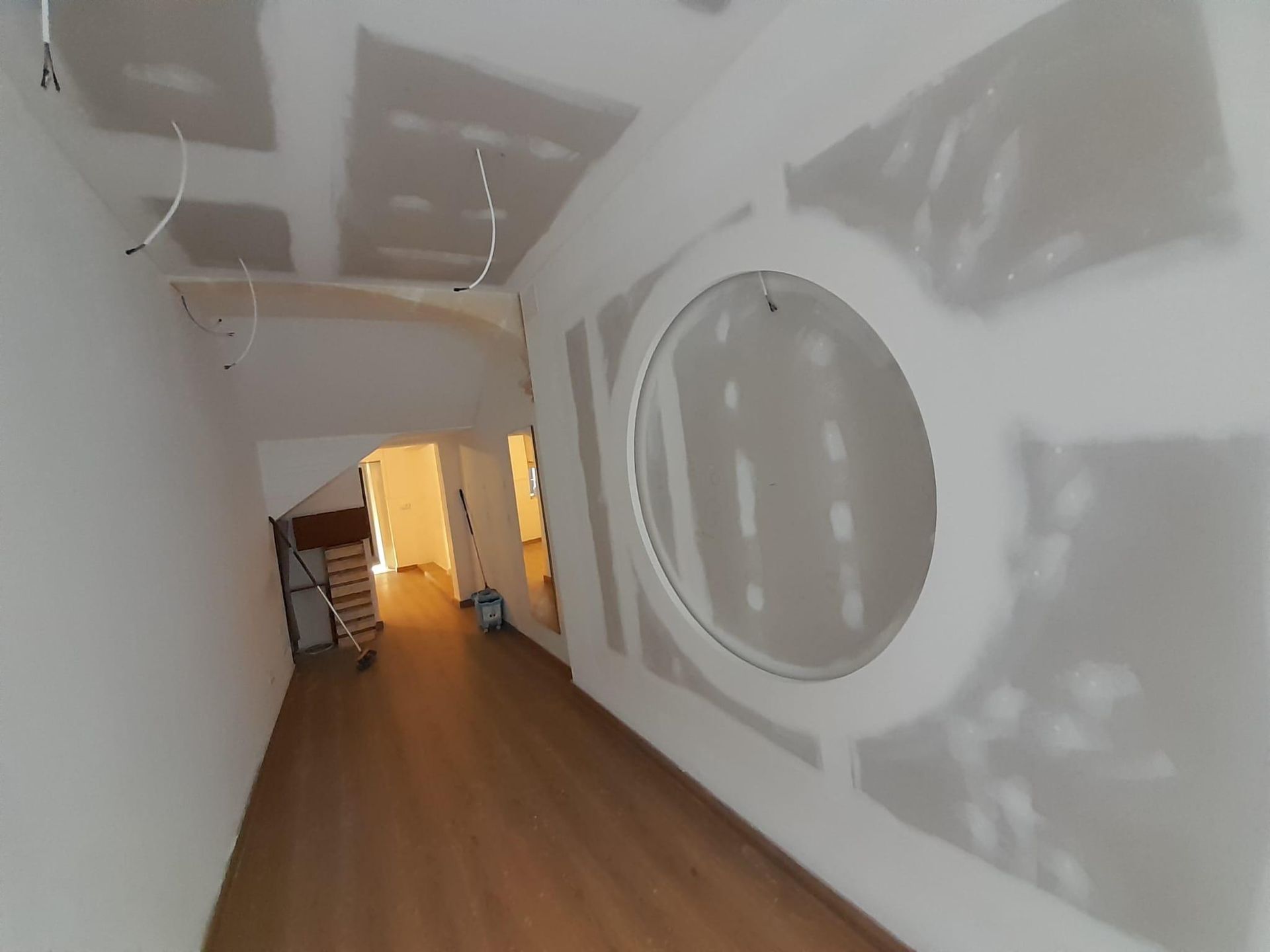 Un pasillo en construcción con paredes de pladur blancas, un gran hueco circular en la pared derecha y suelo de madera.