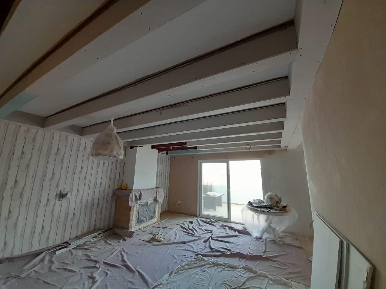 Una habitación en proceso de renovación con vigas de techo a la vista, paredes parcialmente cubiertas con papel pintado