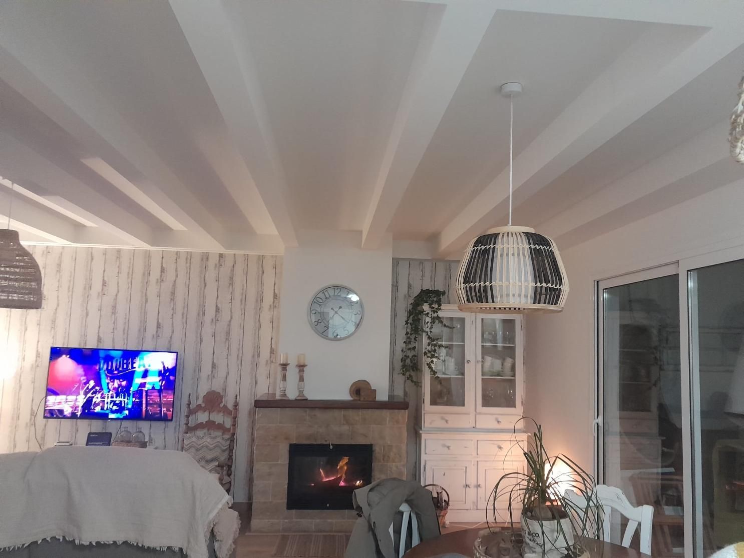 Sala de estar con chimenea, televisor en una pared estampada, un armario blanco y una lámpara colgante de mimbre.