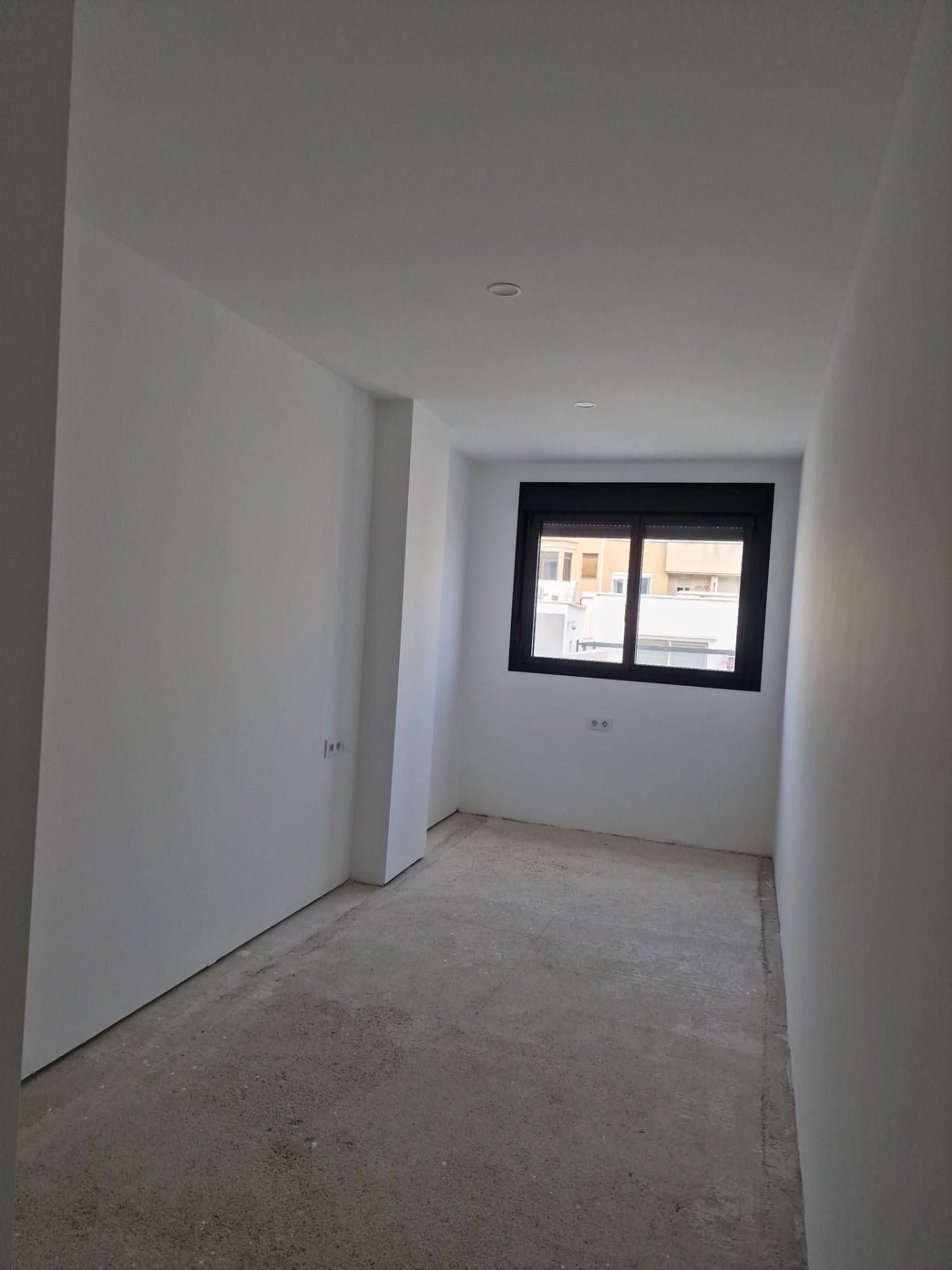 Una habitación estrecha y vacía, con paredes blancas lisas, una sola ventana con marco negro