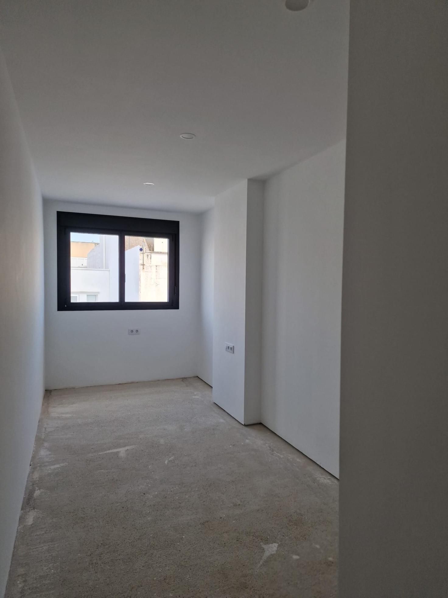 Una habitación vacía y minimalista con paredes blancas, una ventana con marco oscuro y suelo de hormigón sin acabado.