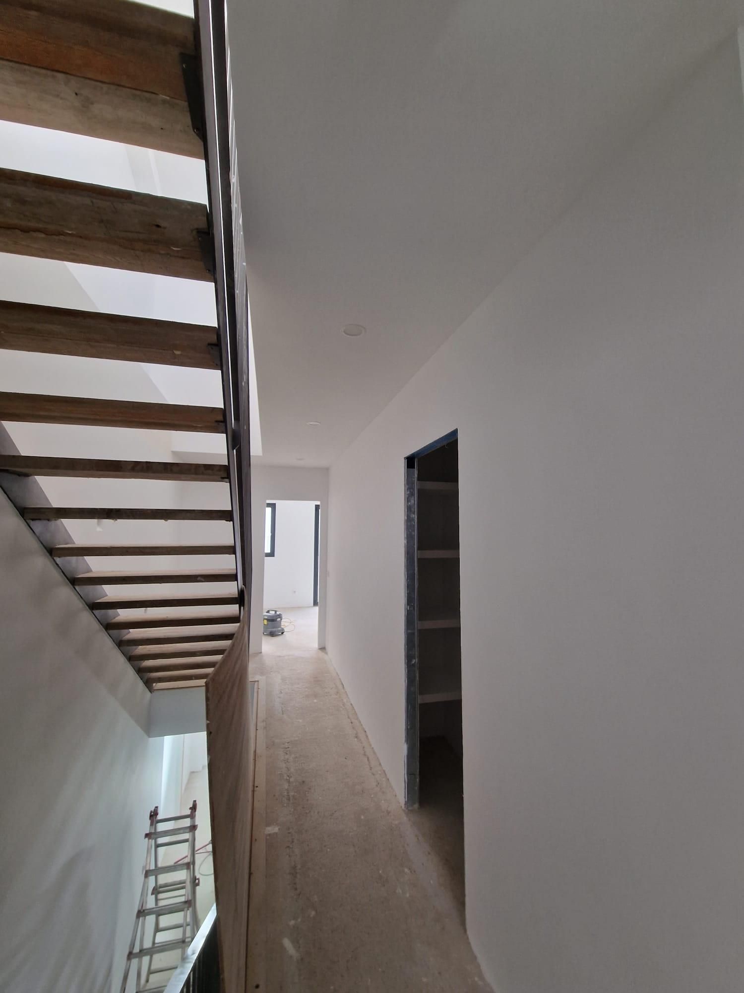 Un pasillo en una casa en construcción con una escalera de madera a la izquierda, paredes blancas y vista a otra habitación.