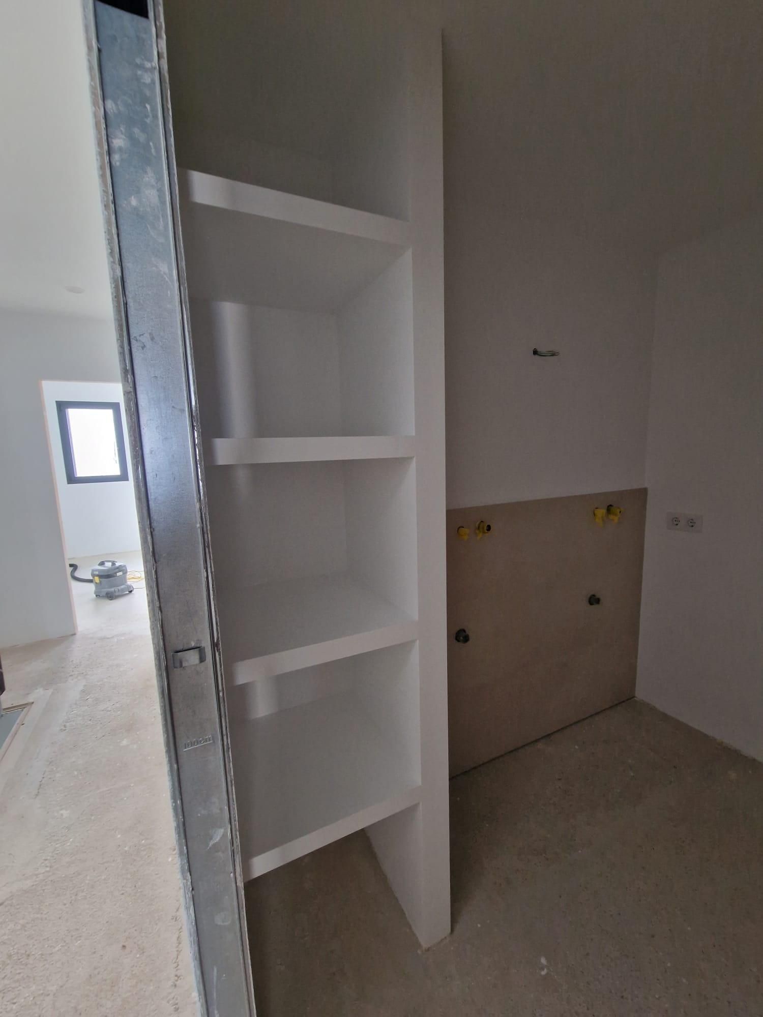 Una habitación sin terminar que cuenta con una estantería blanca empotrada junto a una pared con paneles de yeso a la vista.