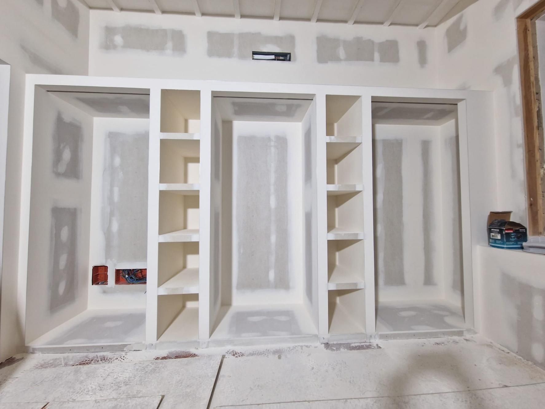 Una pared en construcción con tres armarios empotrados con estantes, acabada en paneles de yeso blancos
