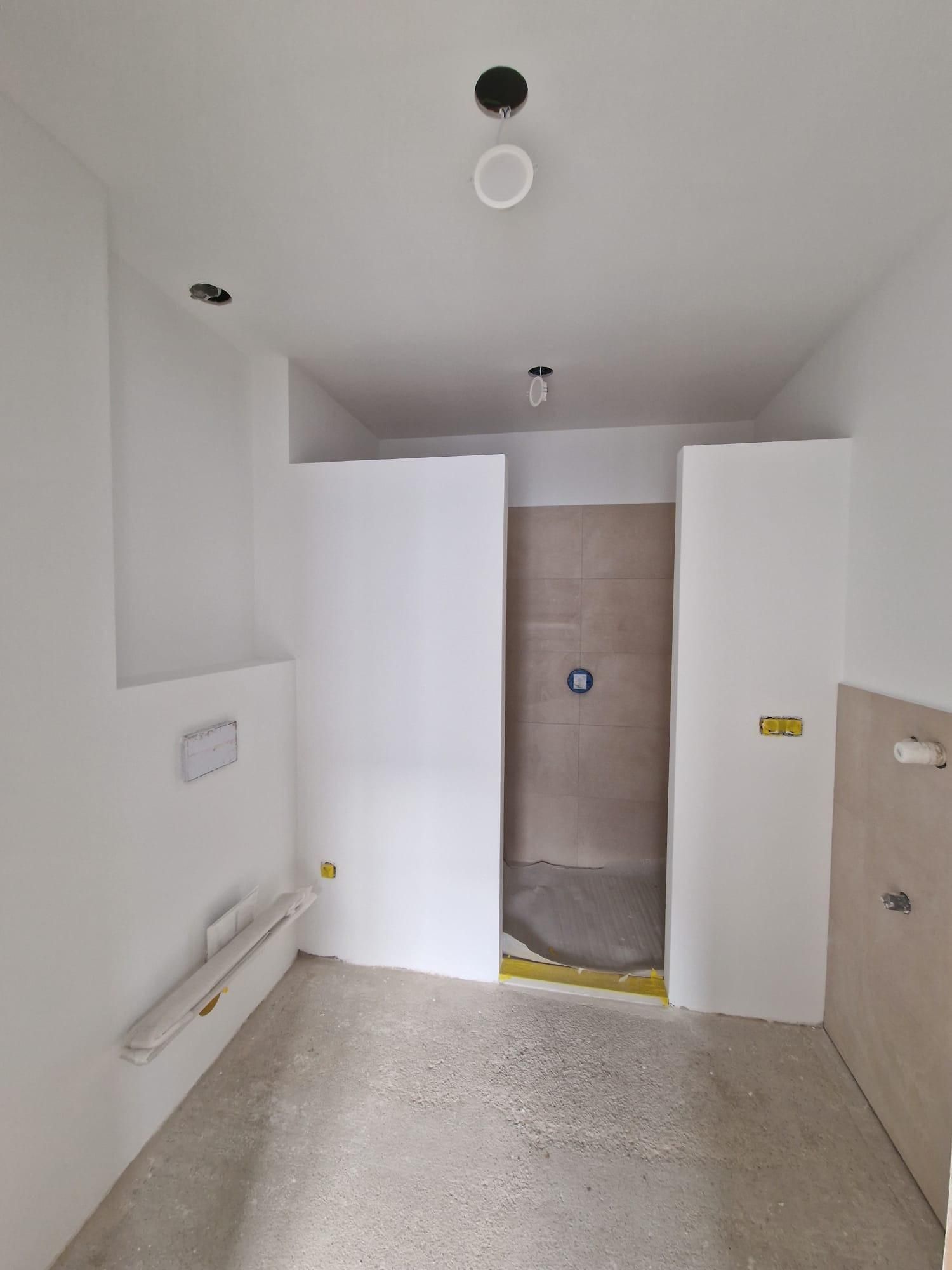 Un baño moderno en construcción con paredes blancas, zona de ducha alicatada y suelos de hormigón sin acabar.