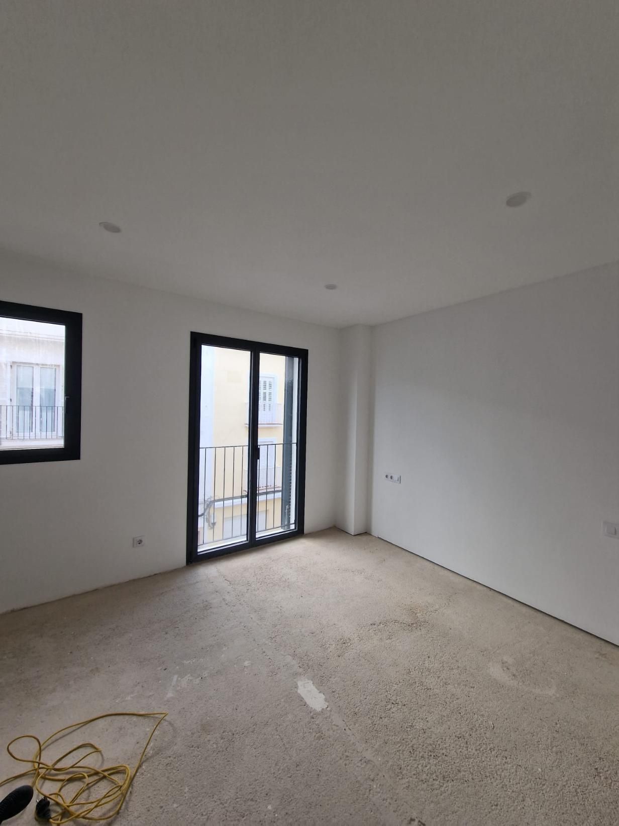 Una habitación vacía y sin amueblar, con paredes blancas, luces empotradas en el techo, ventanas con marcos oscuros