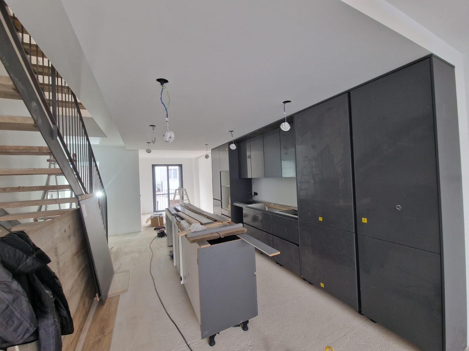 Una cocina en construcción con gabinetes de color gris oscuro, una isla sin terminar y una escalera de madera a la izquierda.