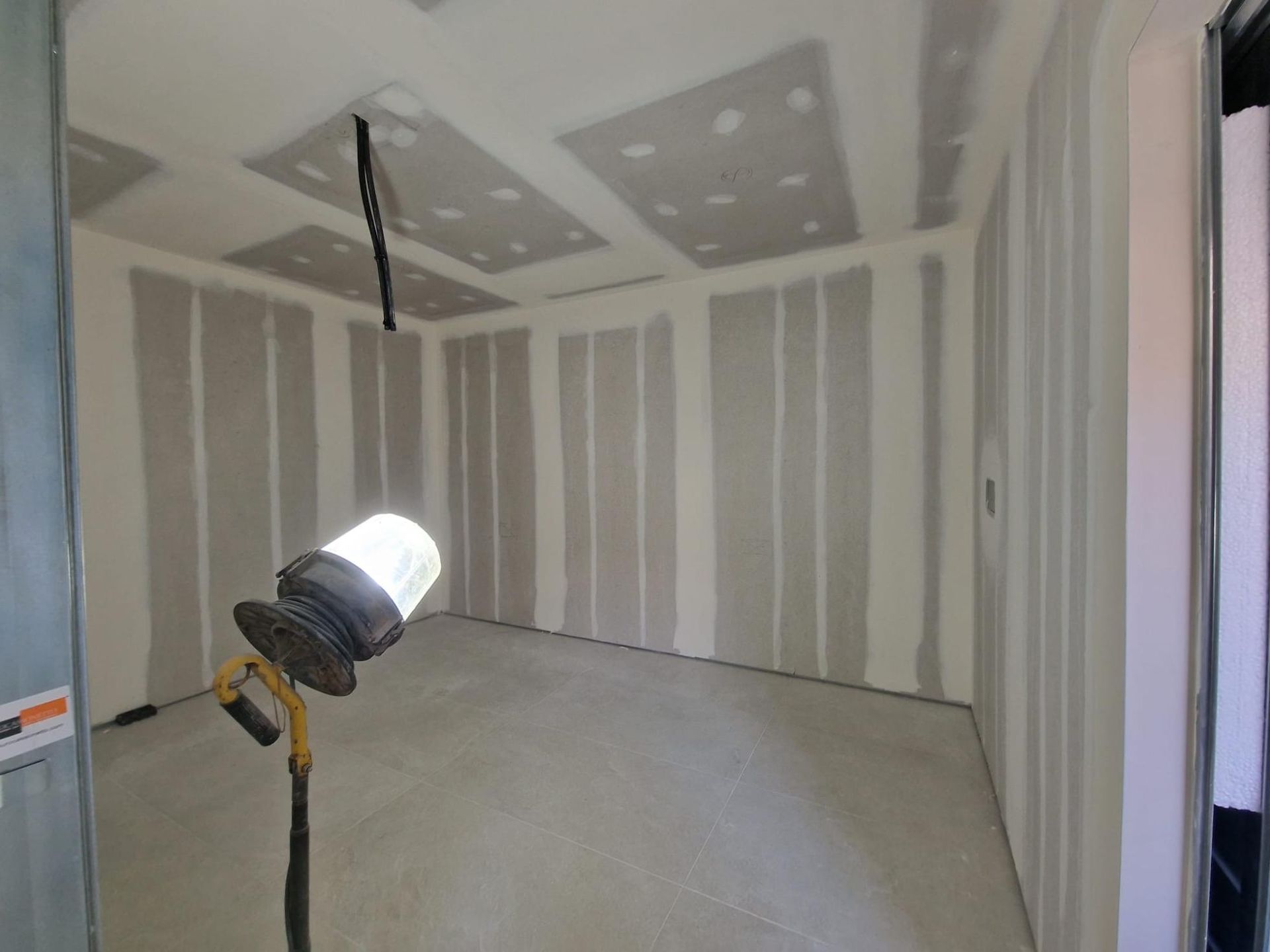 Una lámpara de trabajo ilumina una habitación en construcción con paneles de yeso sin terminar en las paredes y el techo.