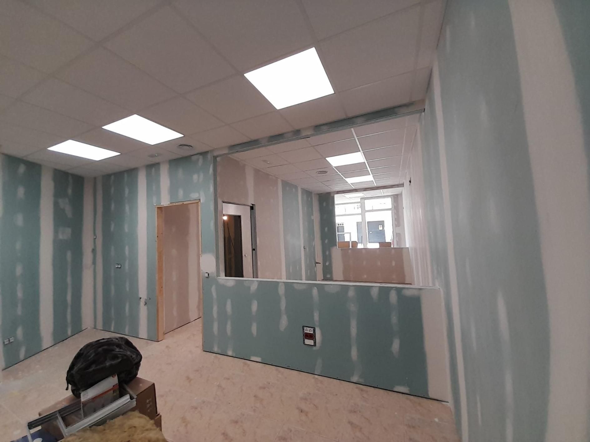 Una oficina en construcción con paneles de yeso de color azul claro, luces empotradas en el techo y una puerta abierta.
