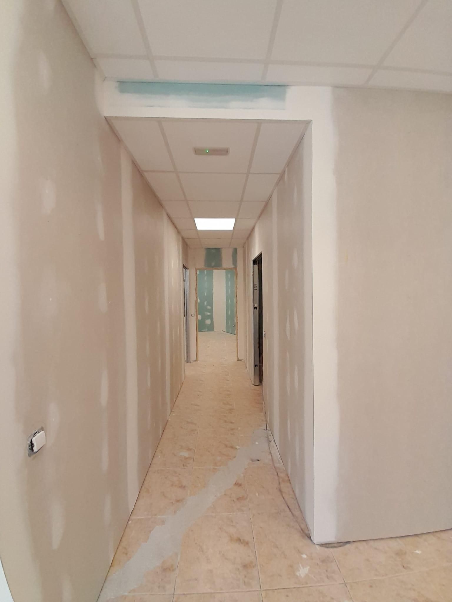 Un pasillo largo y estrecho en construcción, con paredes de pladur sin terminar, un techo suspendido y suelo de color claro.