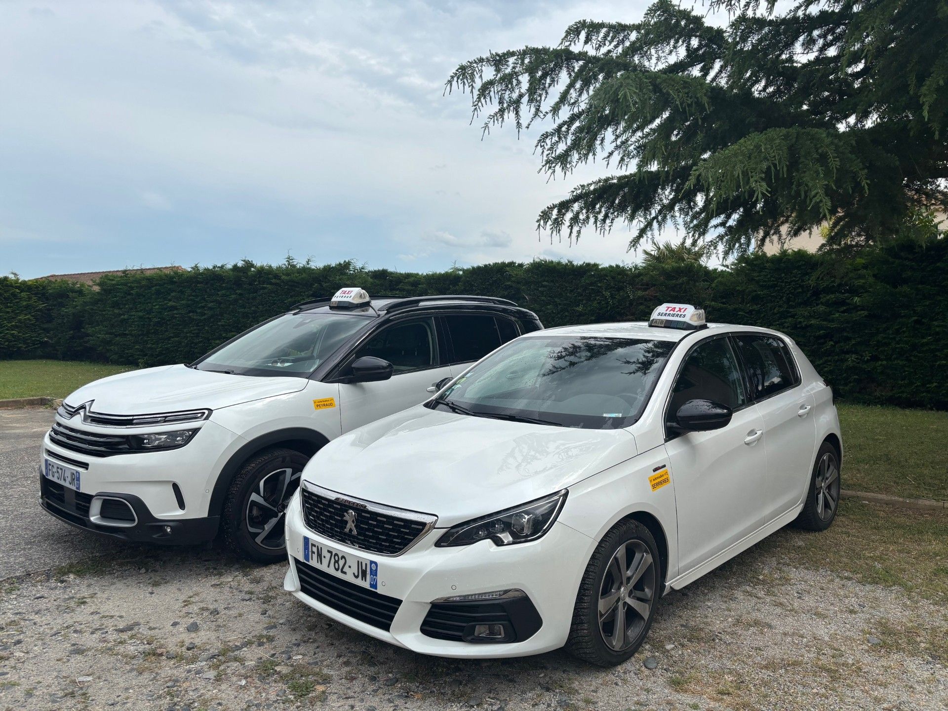 Deux taxis blancs stationnés à l'extérieur, sur une zone gravillonnée, près d'une haie.
