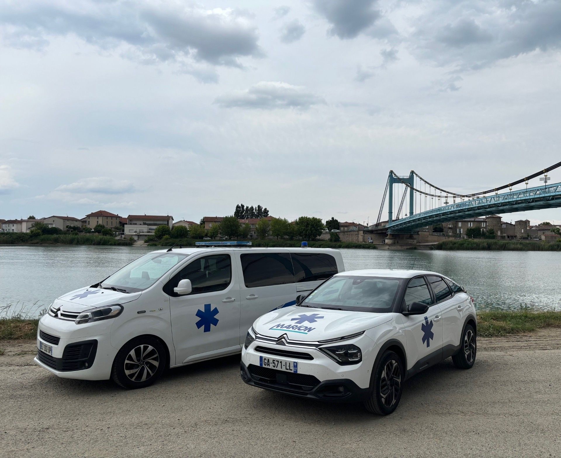 Deux ambulances blanches ornées d'étoiles bleues de vie sont stationnées au bord de l'eau, avec un pont en arrière-plan.