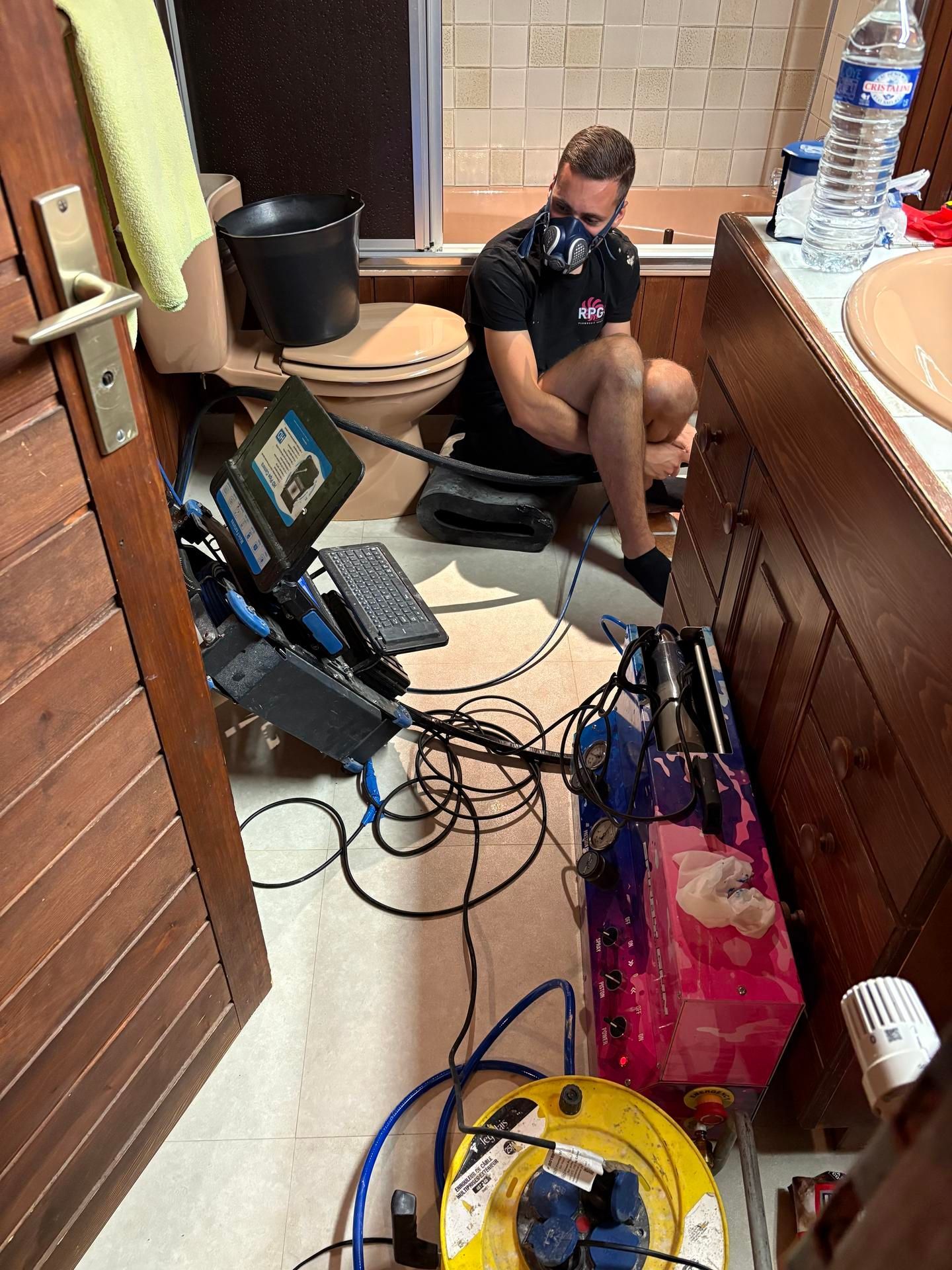 Un homme portant un masque se trouve dans une salle de bains, entouré d'équipements : ordinateur, câbles, multiprise.