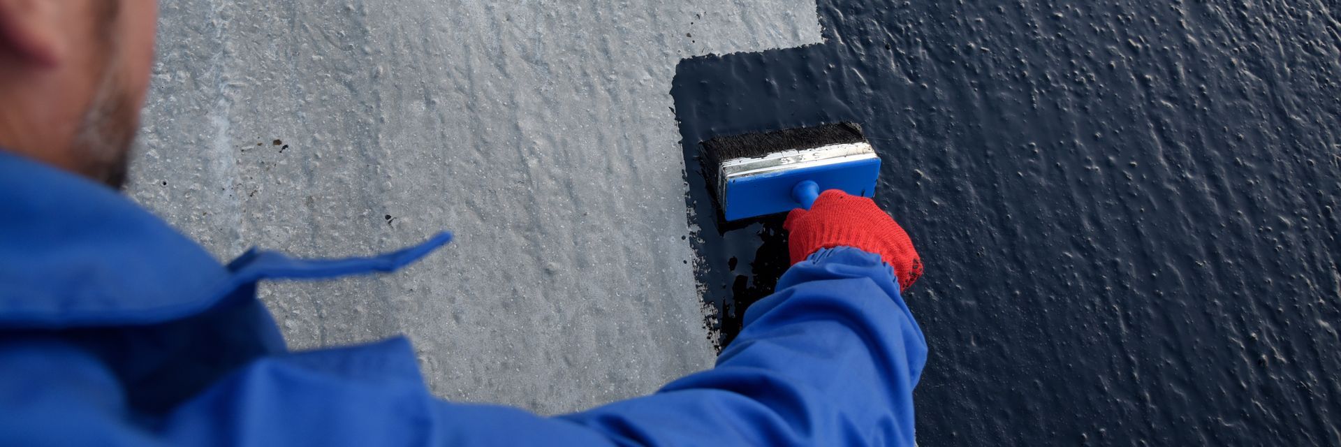 Personne appliquant un mastic noir sur une surface à l'aide d'un pinceau ; portant une combinaison bleue et des gants rouges.