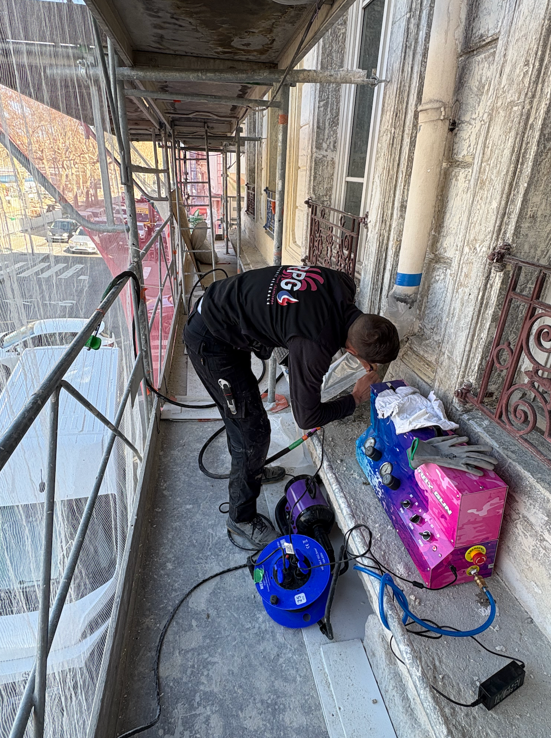 Un ouvrier répare la façade d'un immeuble depuis un balcon ; des échafaudages entourent le bâtiment.