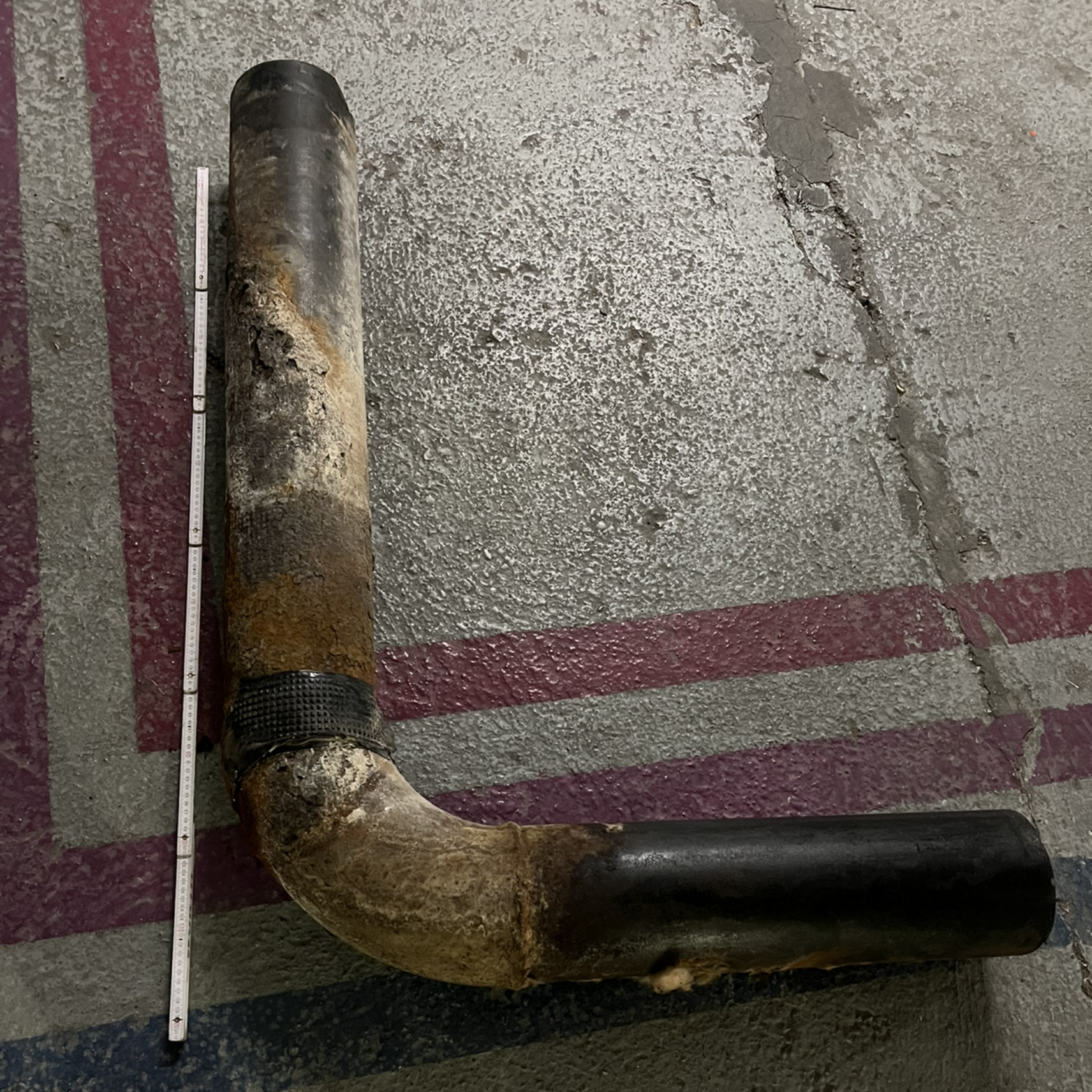 Tuyau en forme de L, corrodé, posé sur un sol en béton. Une règle graduée est placée à côté du tuyau.