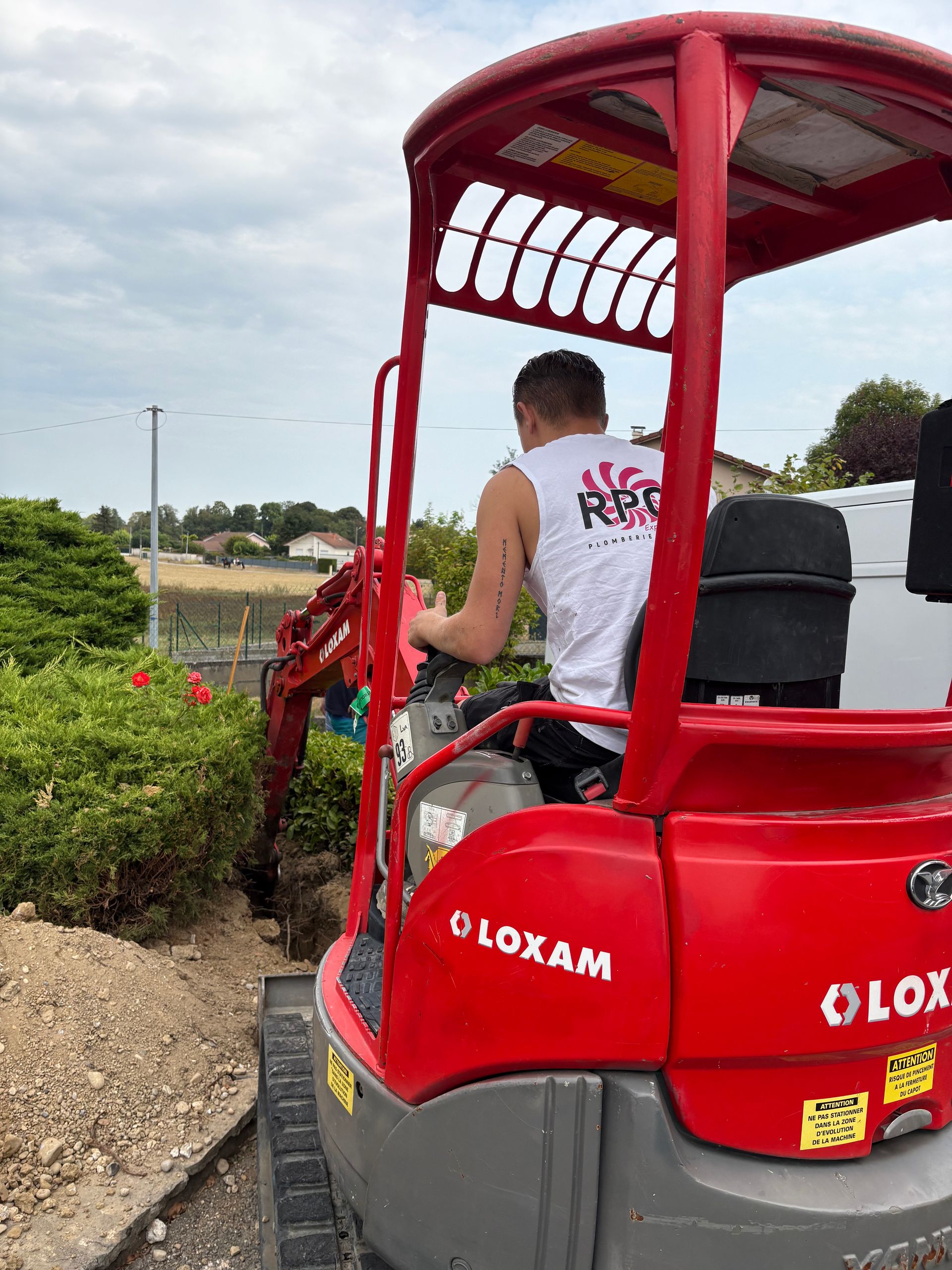 Un homme manœuvre une pelle mécanique LOXAM rouge et creuse près de buissons.