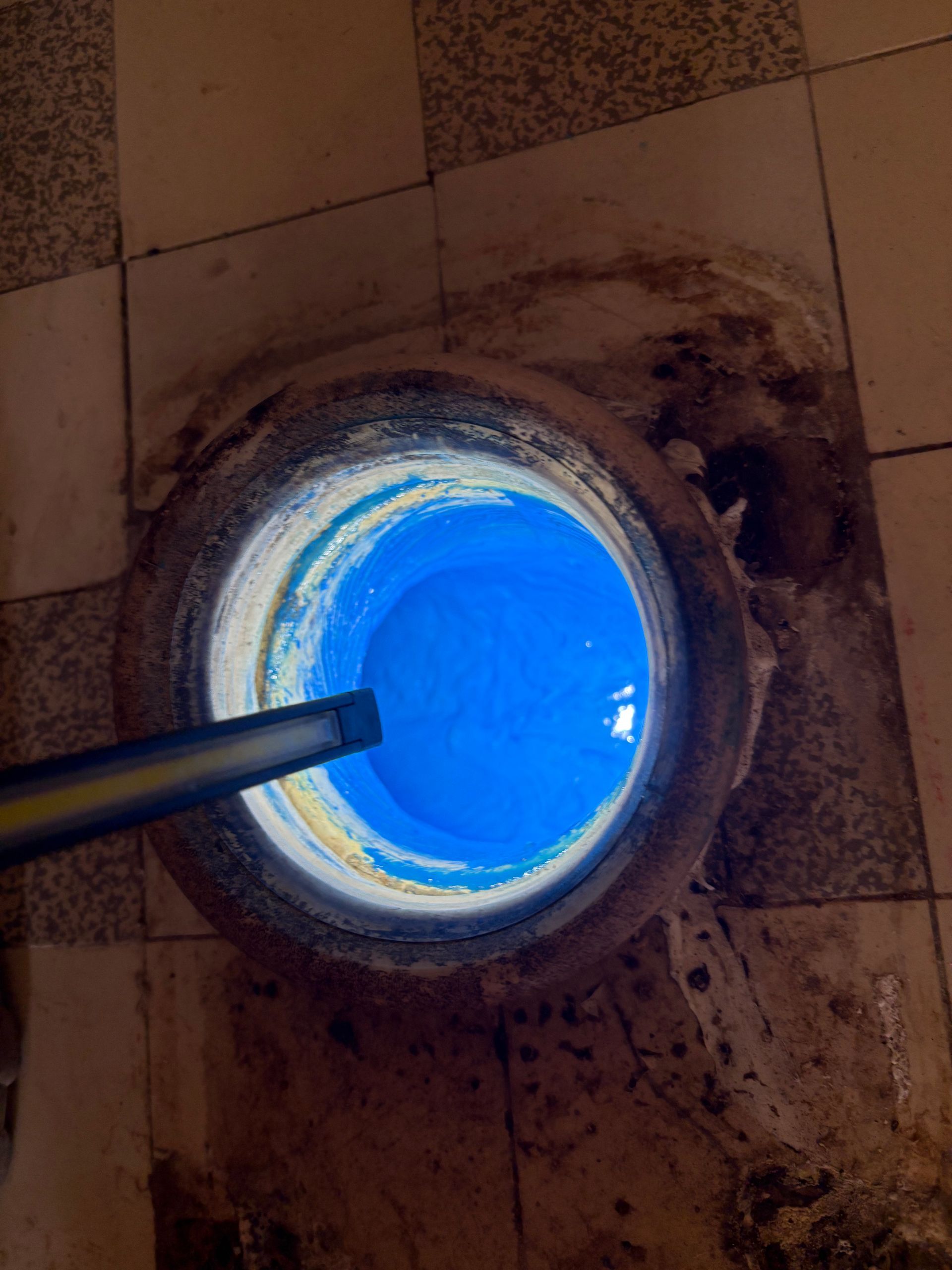Vue aérienne d'un puits circulaire rempli de liquide bleu, posé sur un sol carrelé et contenant un tuyau d'arrosage.