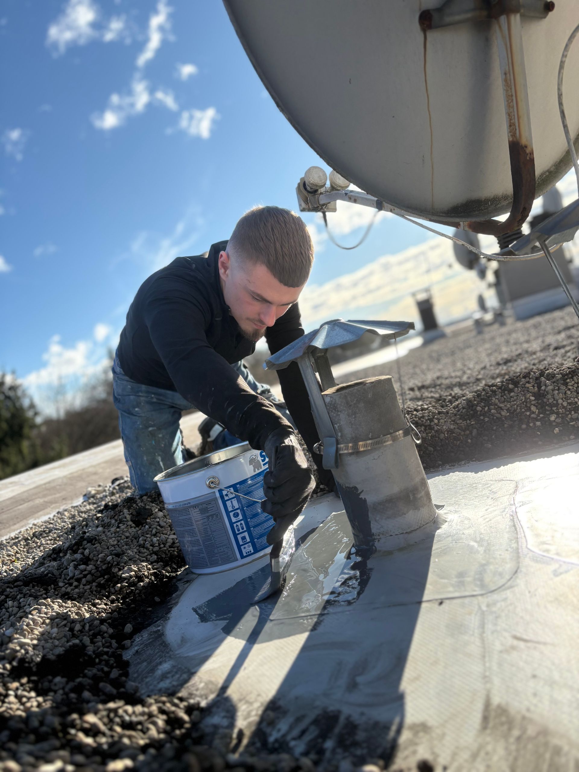 Une personne, perchée sur un toit, applique du mastic sur une structure, à côté d'une antenne parabolique.