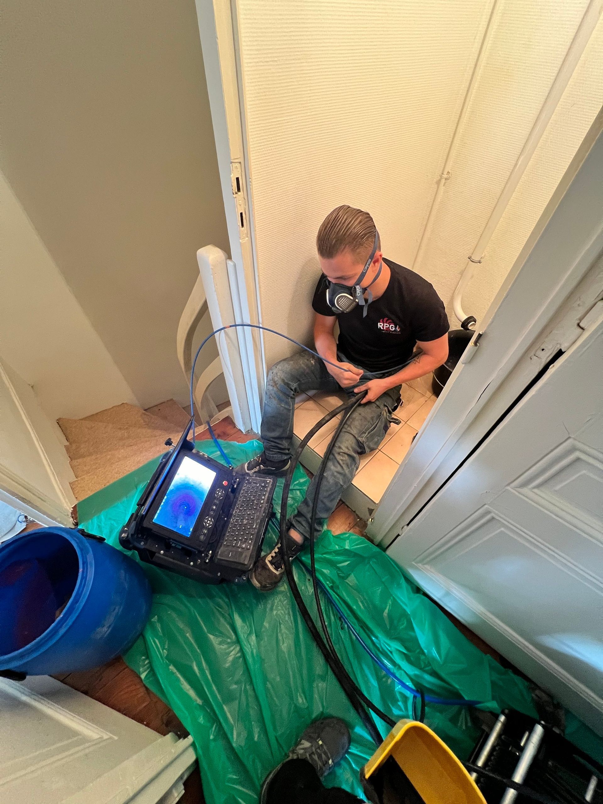 Homme utilisant du matériel, assis sur des marches, bâche verte, seau bleu, murs blancs.