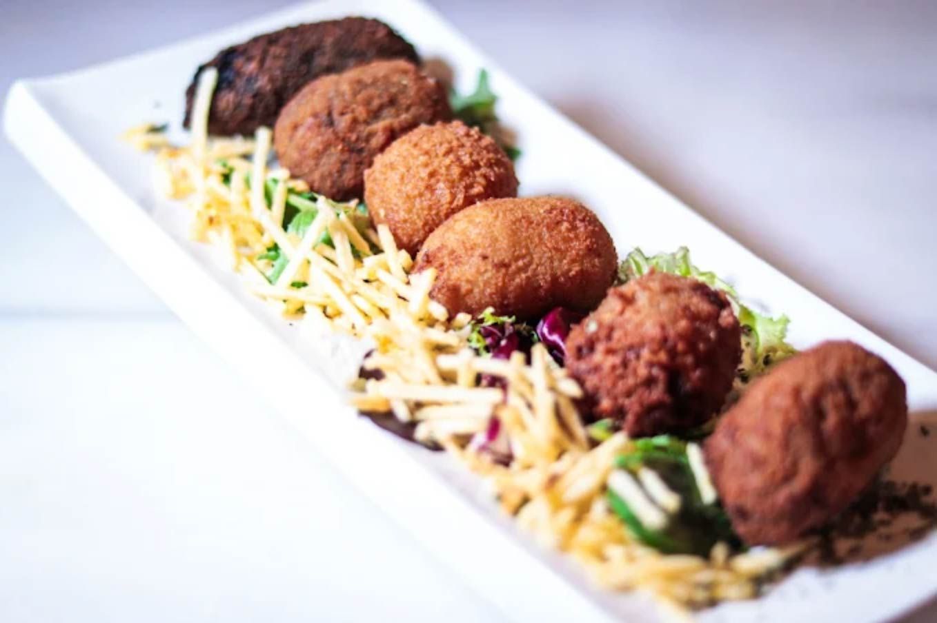 Un plato rectangular blanco con seis aperitivos fritos y redondos sobre patatas ralladas y verduras.
