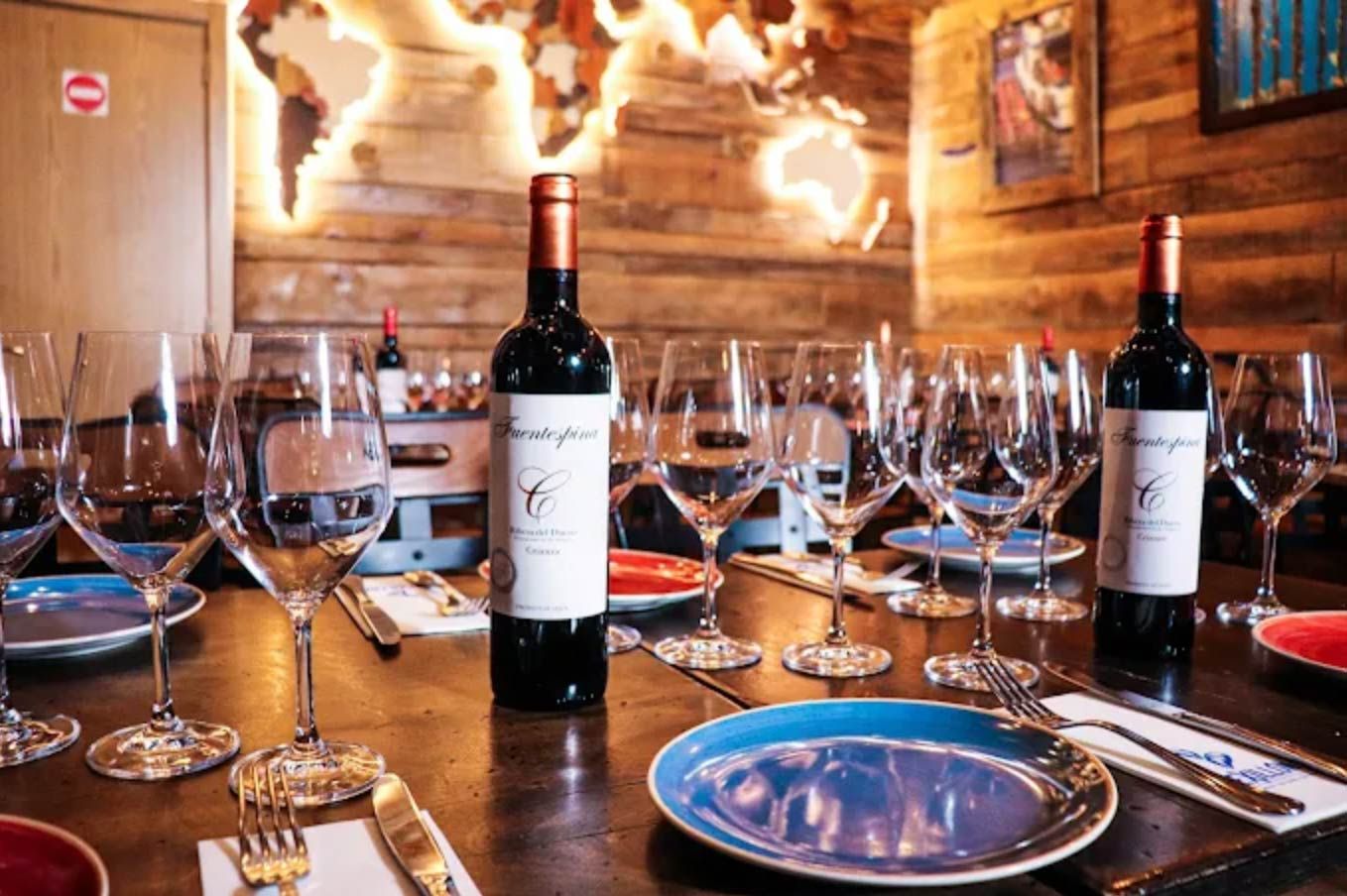 Mesa preparada para cata de vinos con botellas, copas y platos. Pared rústica de madera con mapamundi.