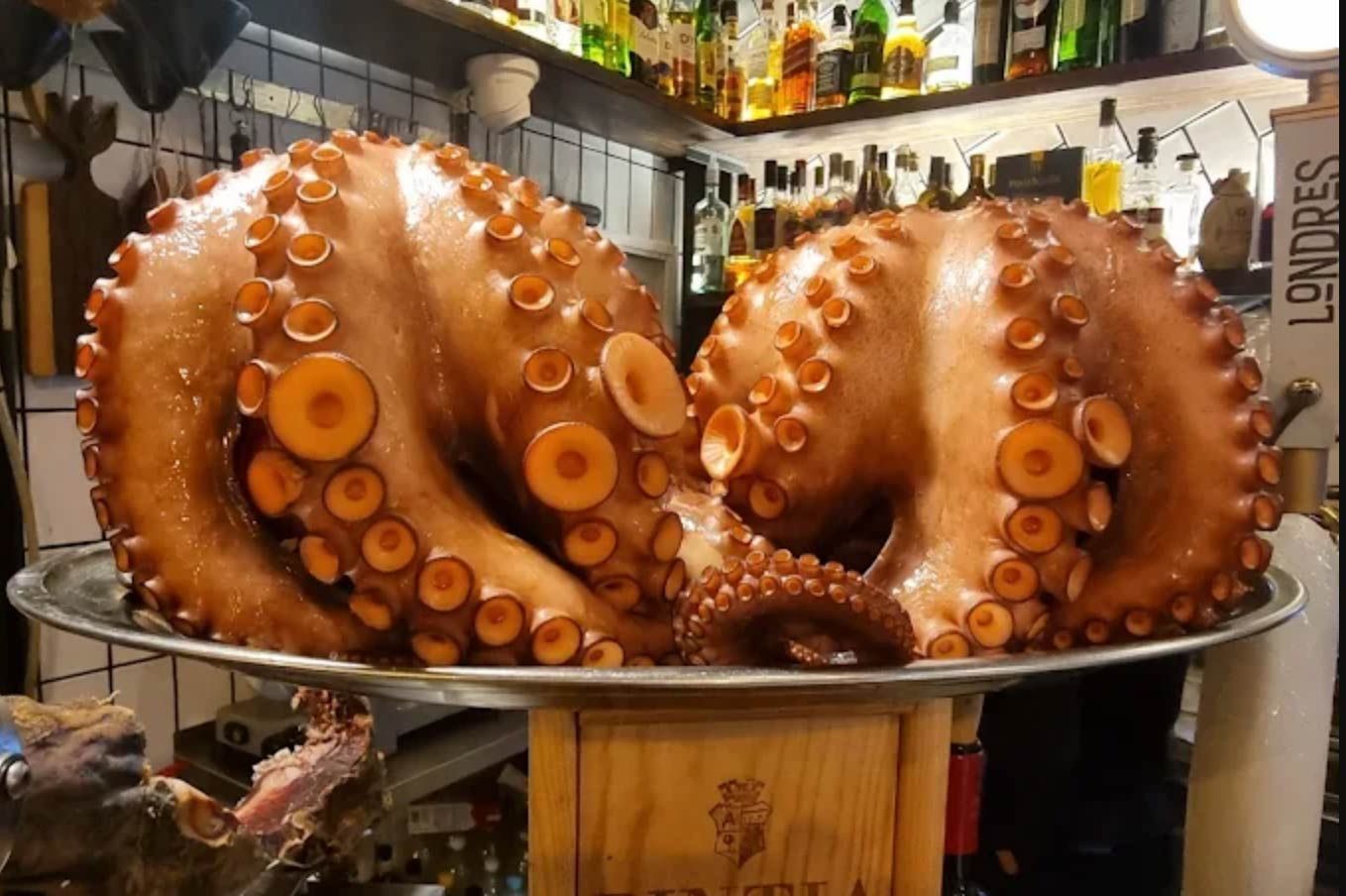 Dos pulpos cocidos en bandeja de plata, en un restaurante, con botellas al fondo.