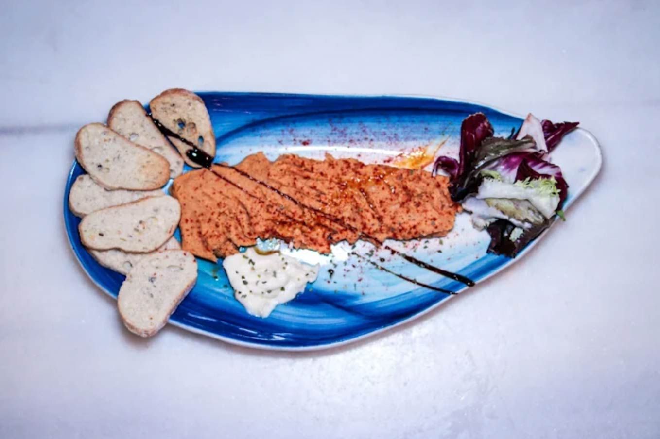 Aperitivo en plato azul: salsa roja, rebanadas de pan, verduras y un chorrito de salsa.