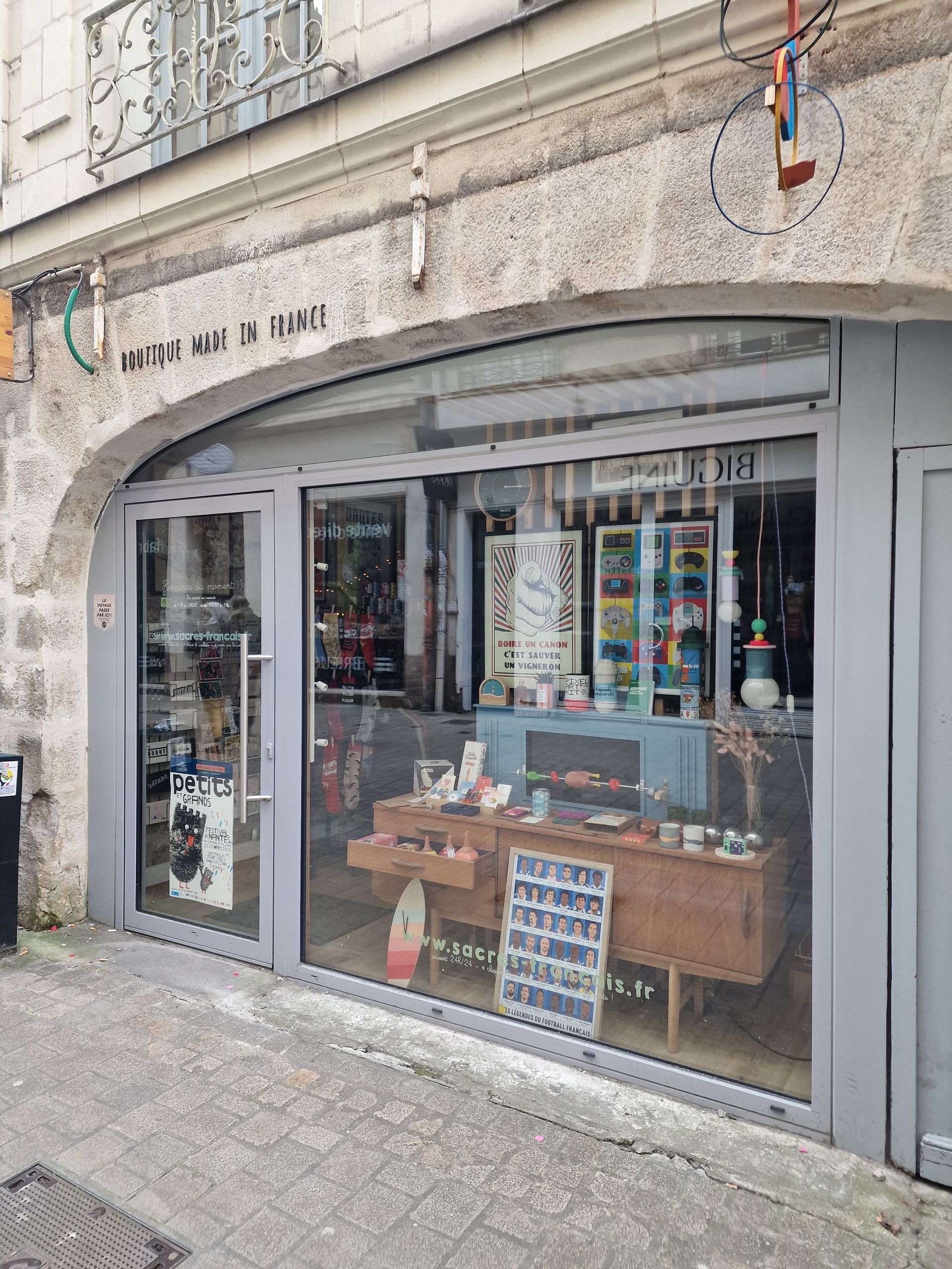 Façade de magasin avec de grandes vitrines exposant divers articles. Façade en pierre grise et porte cintrée.