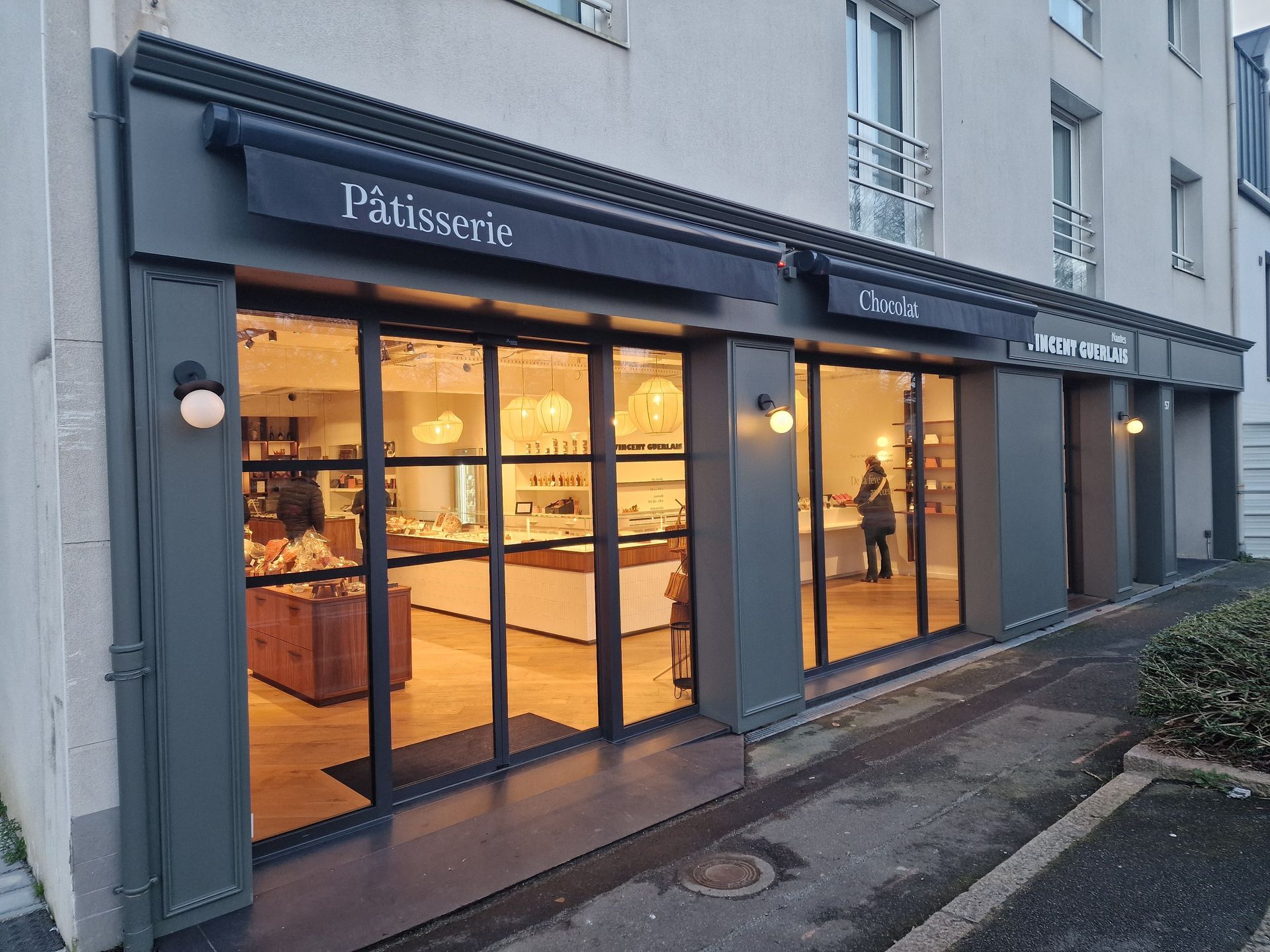 Façade de boulangerie avec de grandes vitrines et des auvents noirs ; intérieur éclairé présentant des pâtisseries.