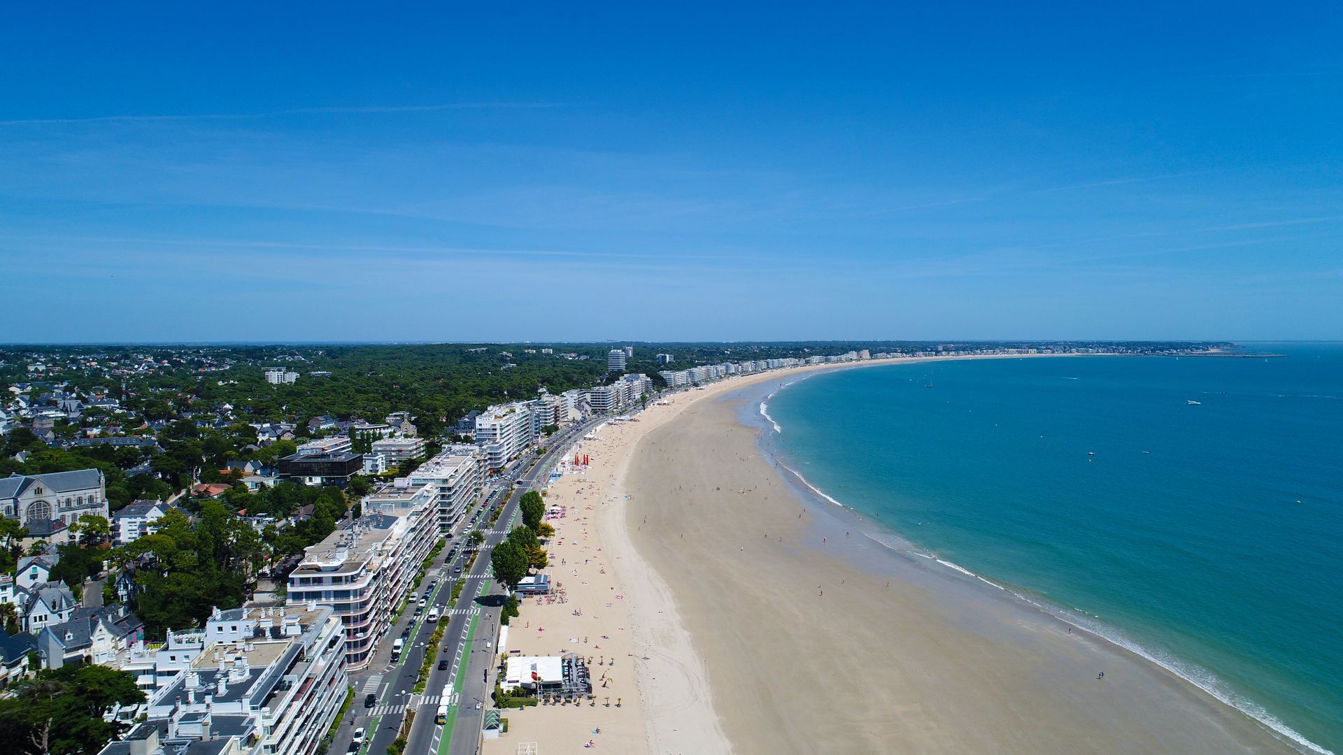 Vue aérienne d'une plage de sable fin, d'un océan turquoise et d'une ville côtière avec des bâtiments et des arbres.