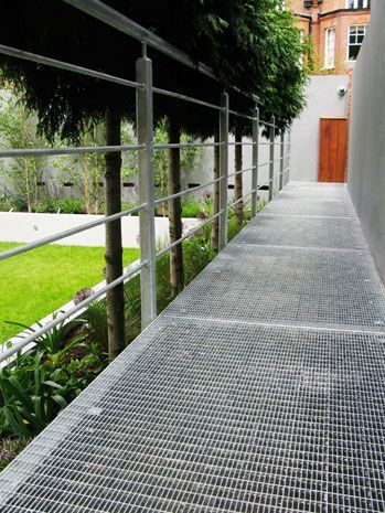 Passarela metálica com corrimão ao lado de um gramado bem cuidado, árvores e um prédio.