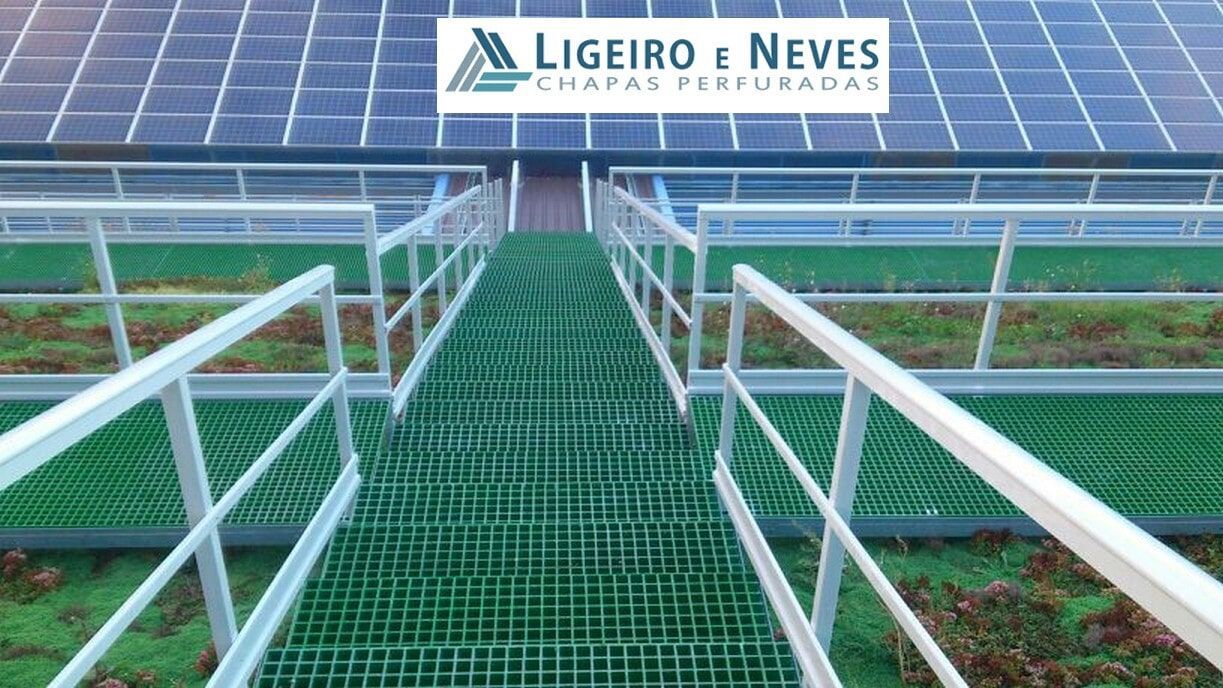 Passarela metálica perfurada verde com corrimãos brancos, que leva a painéis solares no telhado.