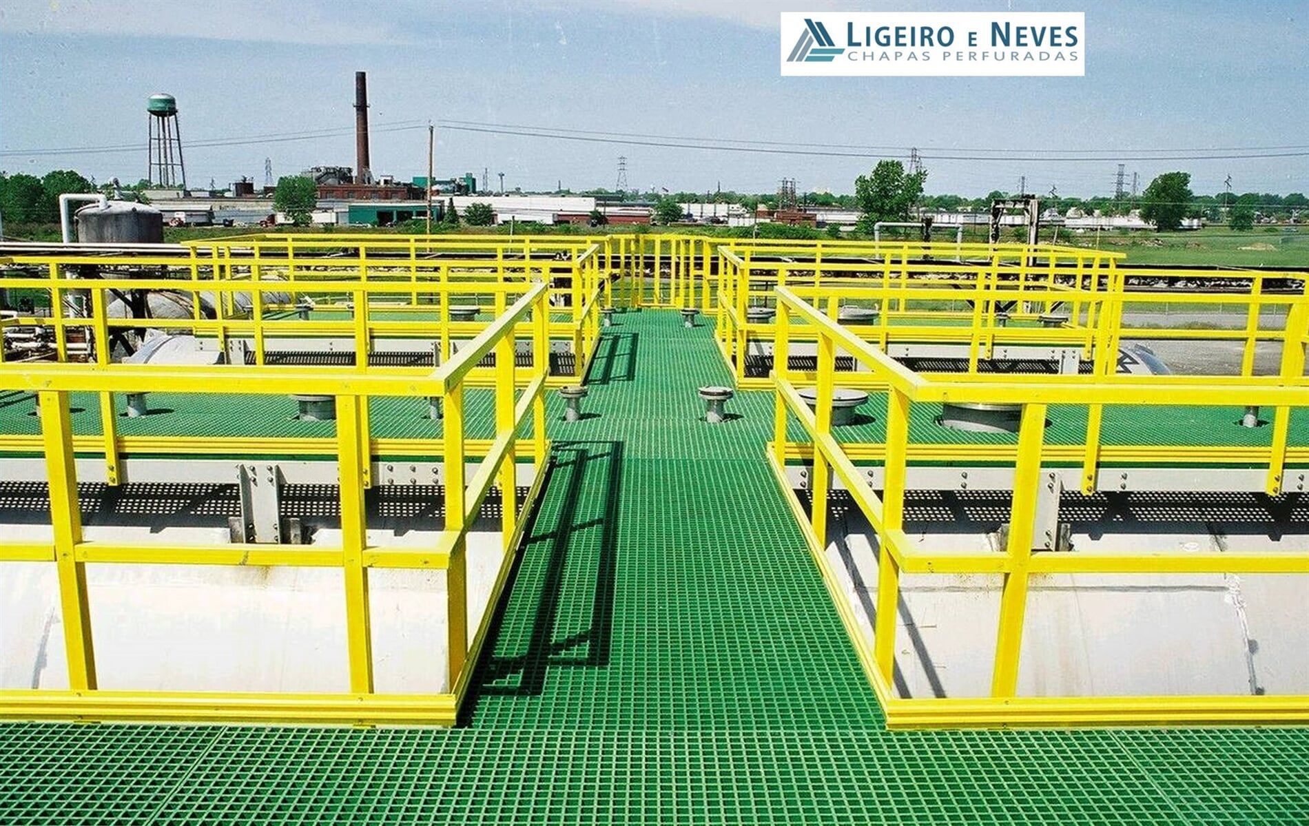 Passarela gradeada verde com corrimãos amarelos em um ambiente industrial.