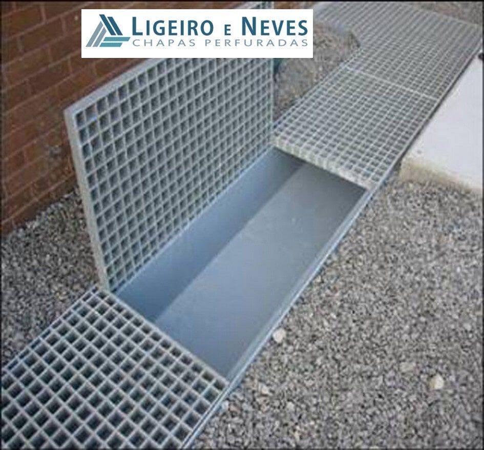 Ralo metálico aberto com tampa. Cinza, embutido no cascalho, próximo a uma parede de tijolos.