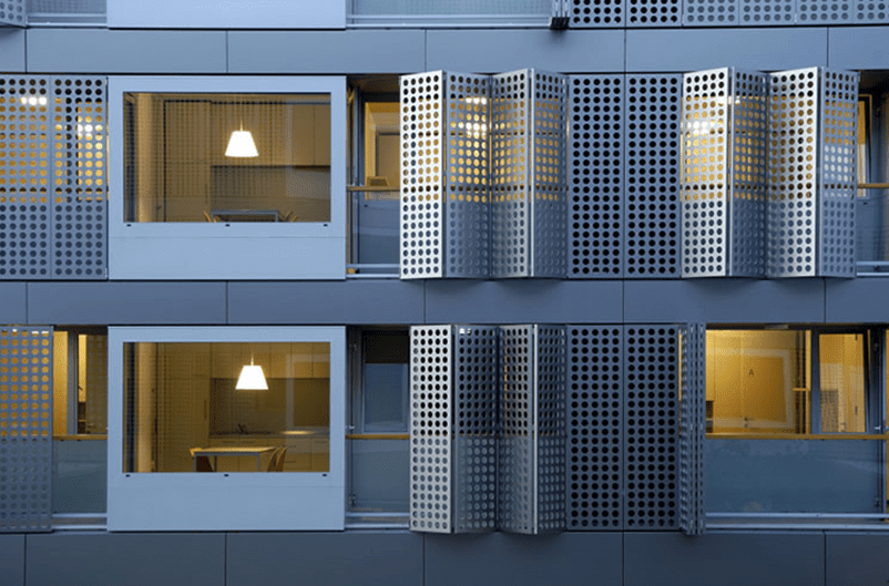 Fachada de edifício moderno com persianas quadradas de metal parcialmente abertas, revelando interiores iluminados.