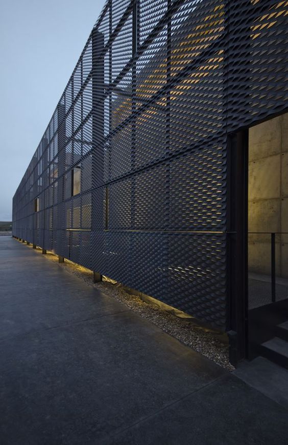 Fachada de um edifício em malha metálica cinza-escura com uma porta aberta e iluminação exterior.